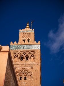 The Koutoubia minaret rises against a deep blue Marrakesh sky, its carved arches, geometric motifs, turquoise tile band and golden finial glowing in crisp light. Even a small cloud and a perched bird are perfectly visible. Every element is sharp. Download the full HD version to reveal exquisite details etched into centuries old stone.