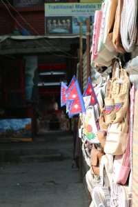 A picture focused on four flags of Nepal hanging together on thin wooden sticks. They are displayed outside a shop possibly as souvenirs. There are various handmade bags and woven items in neutral tone suggesting a tourist market area. The background is dark and blurred reflecting some kind of shop.